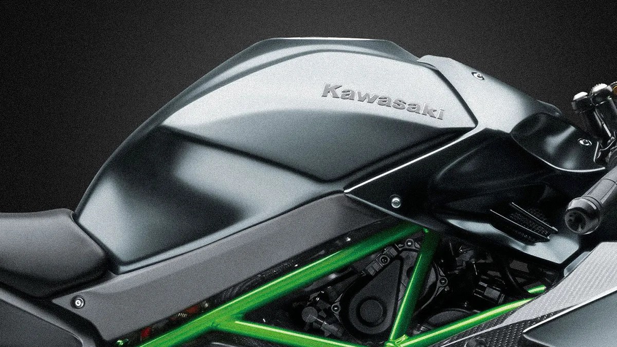 Close-up of a black Kawasaki motorcycle fuel tank with a green frame visible underneath. The Kawasaki logo is embossed on the tank. The image also shows part of the seat and handlebar grip.
