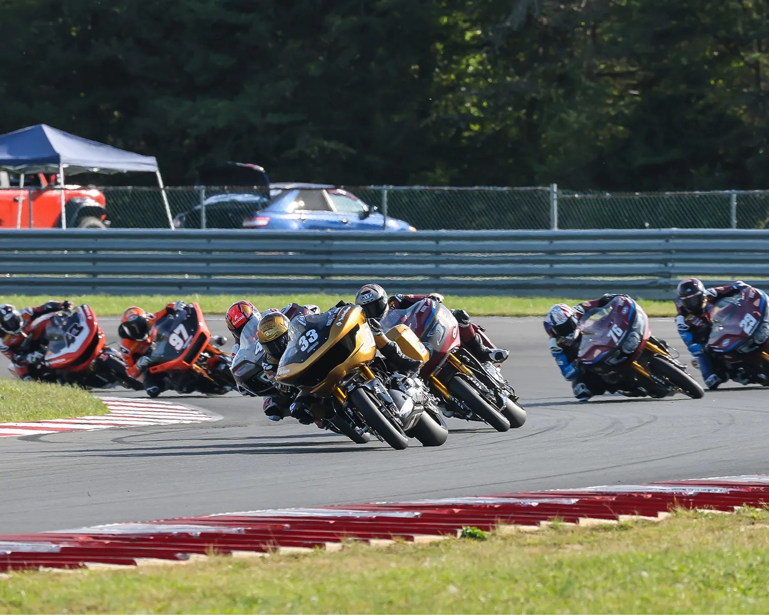 A group of motorcycle racers leaning sharply as they navigate a curve on a racetrack. The leading bike is gold with the number 33, followed closely by a red bike with the number 5. Other racers on various colored bikes trail behind them. The background shows a fence, parked cars, and trees.