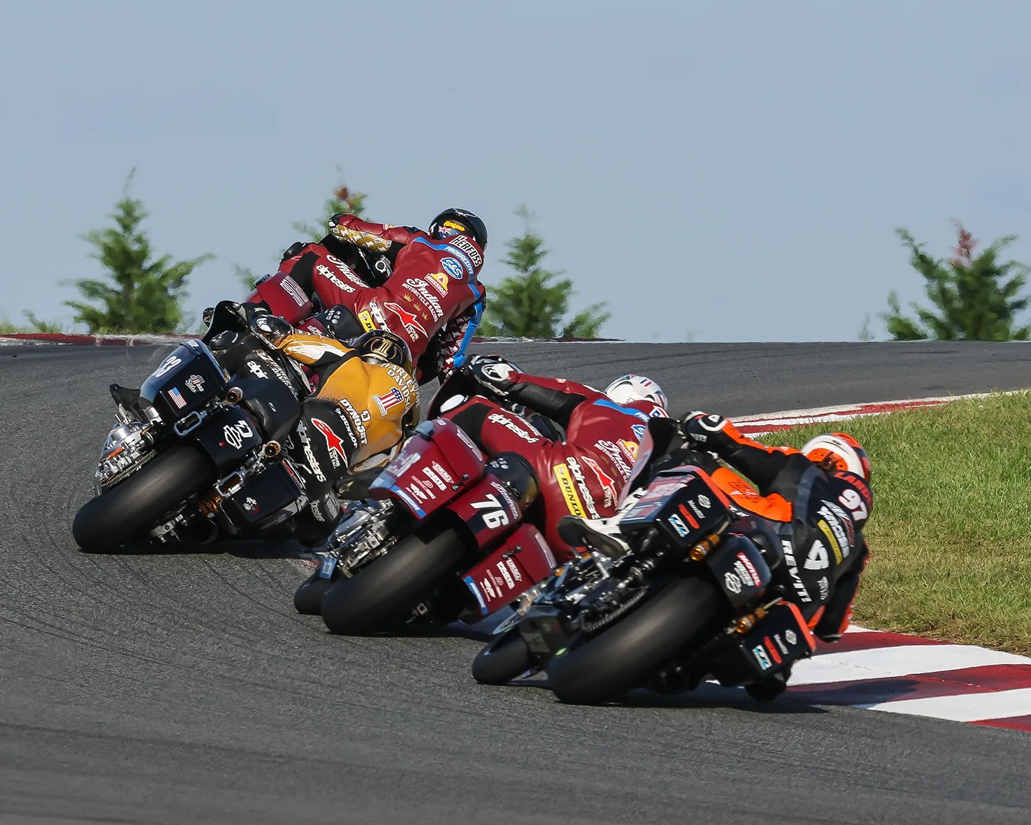 Four motorcycle racers leaning into a sharp turn on a paved race track, with grass on the right side and trees in the background. The riders wear colorful racing suits and helmets, and their bikes display various sponsor logos.