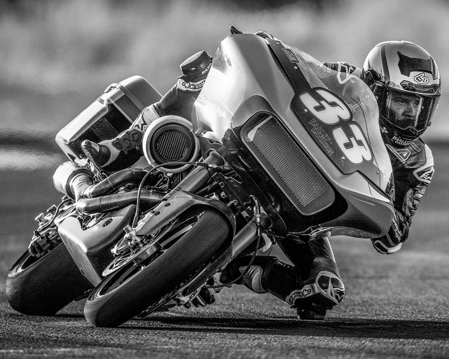 A close-up black and white photo of a motorcycle racer leaning sharply into a turn on a race track. The motorcycle has the number 33 and "Harley Davidson" written on the front fairing. The rider is wearing a full racing suit and helmet, focused intently ahead. The image captures the dynamic motion and intensity of the race.