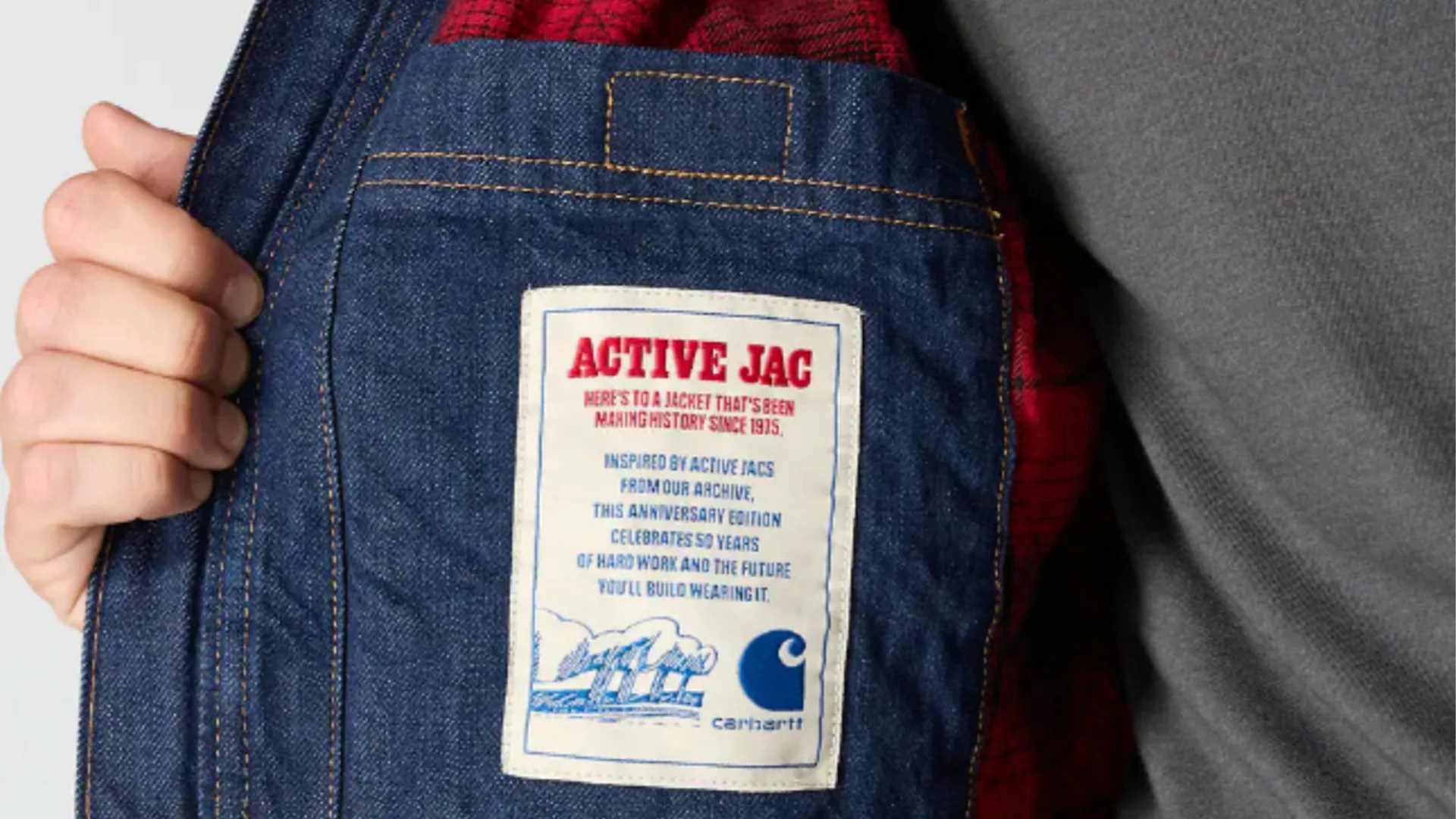 Close-up of the inside pocket of a denim jacket with a white label that reads "ACTIVE JAC" in red, followed by text in blue about the jacket's history and anniversary edition. The label also features a blue Carhartt logo and an illustration of factory smokestacks. The jacket has red and black plaid lining. A hand is holding the jacket open.