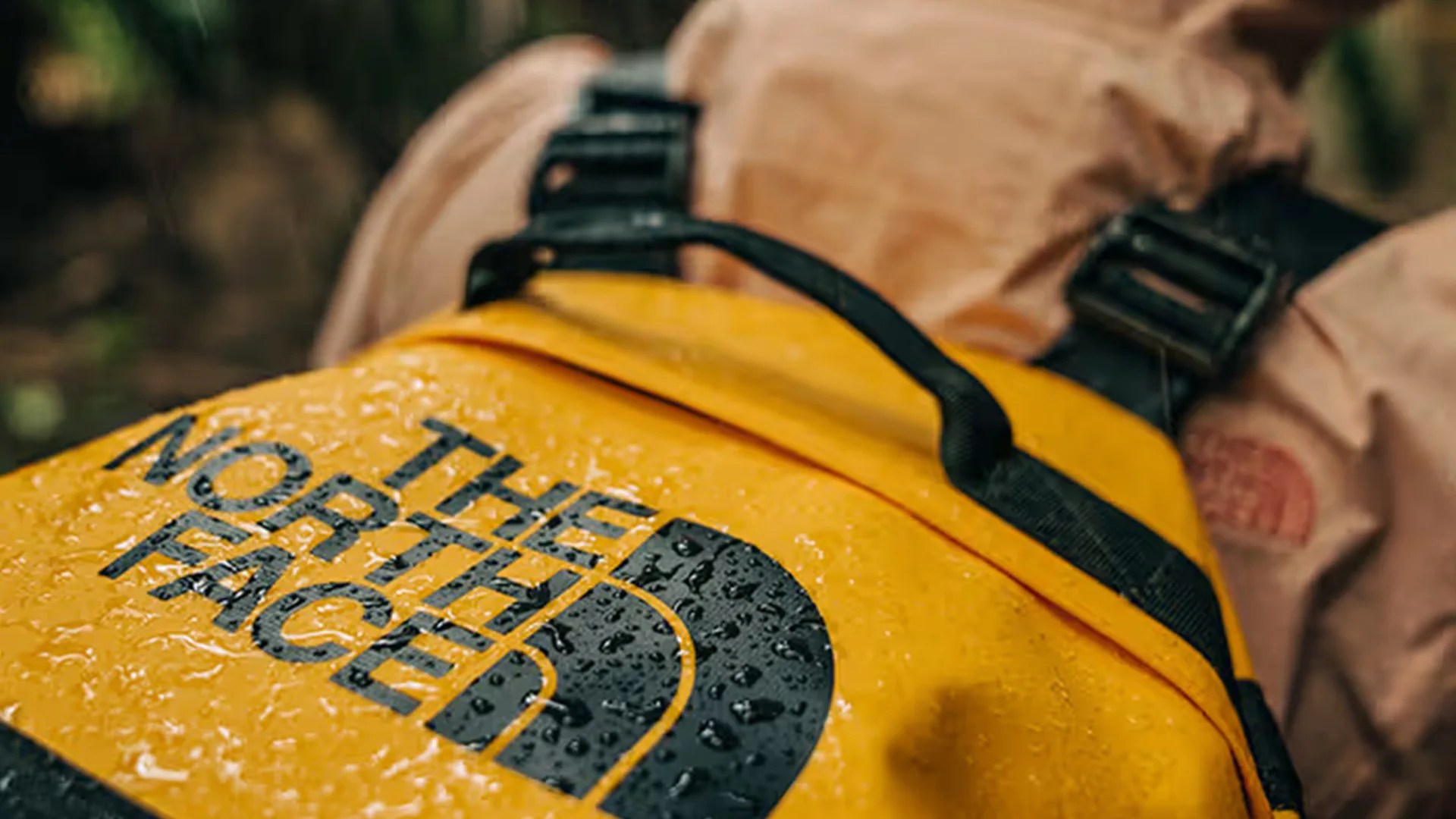 Close-up of a yellow The North Face backpack with water droplets on its surface, featuring black straps and a handle, worn by a person in a light brown jacket.