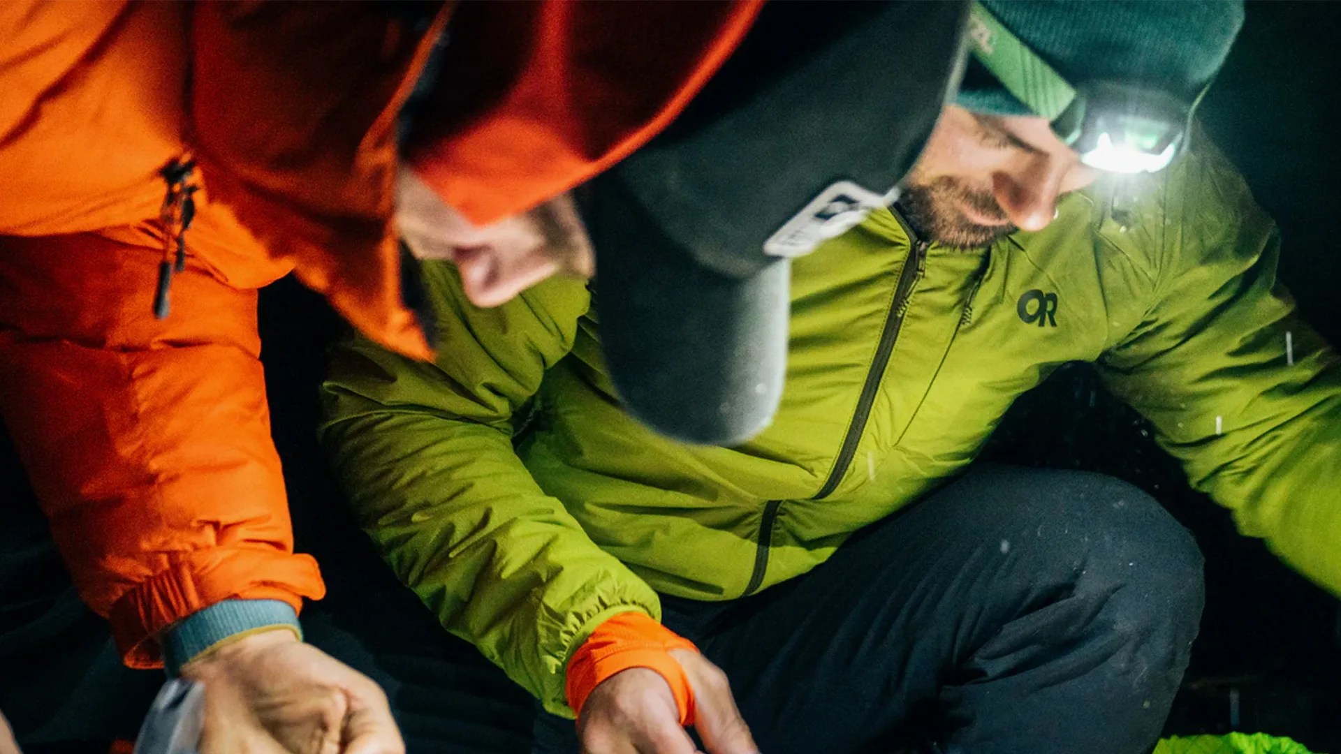 Two people dressed in outdoor gear are closely examining something in low light. One person is wearing an orange jacket, while the other is in a green jacket with a headlamp on their forehead, illuminating the area. Both appear focused and engaged in an activity, possibly camping or hiking at night.