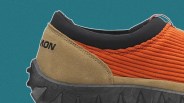 Close-up of the heel and side of a shoe featuring a textured orange fabric upper, tan suede heel panel with black stitching, and a black padded collar. The black rubber sole has a rugged tread pattern. The partial brand name "MON" is visible on the heel.
