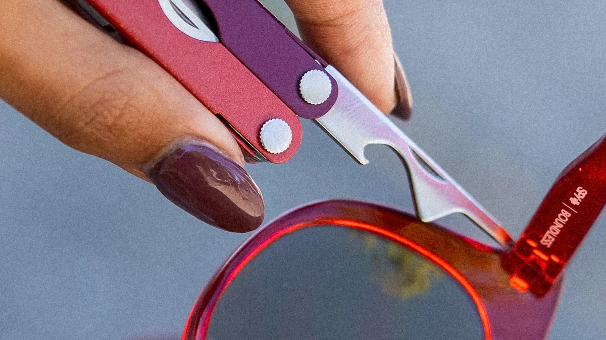 Close-up of a hand with dark purple nail polish using a multi-tool with red and purple handles to adjust or repair the hinge of red-framed sunglasses. The multi-tool features a metal bottle opener or similar tool. The sunglasses have dark lenses and a visible brand name on the arm.