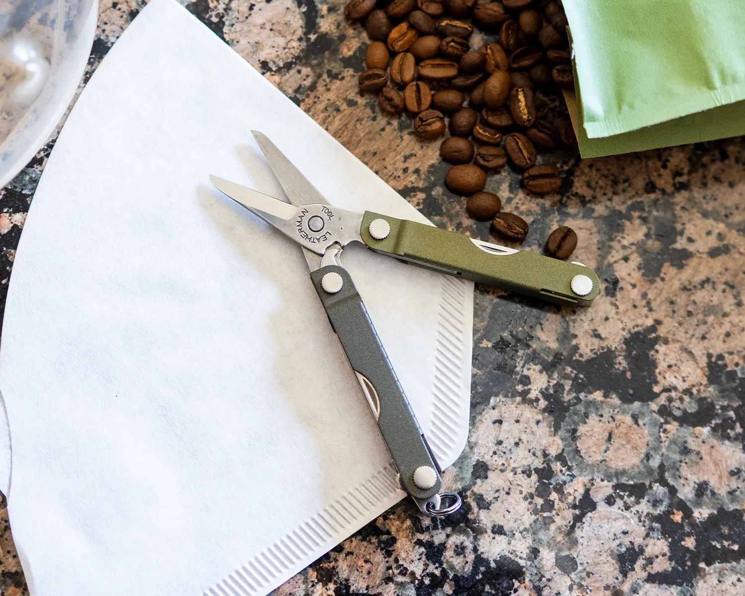 A pair of small, green-handled Leatherman scissors is open and resting on a white coffee filter. Nearby, there are scattered coffee beans and a folded green napkin on a speckled granite countertop.