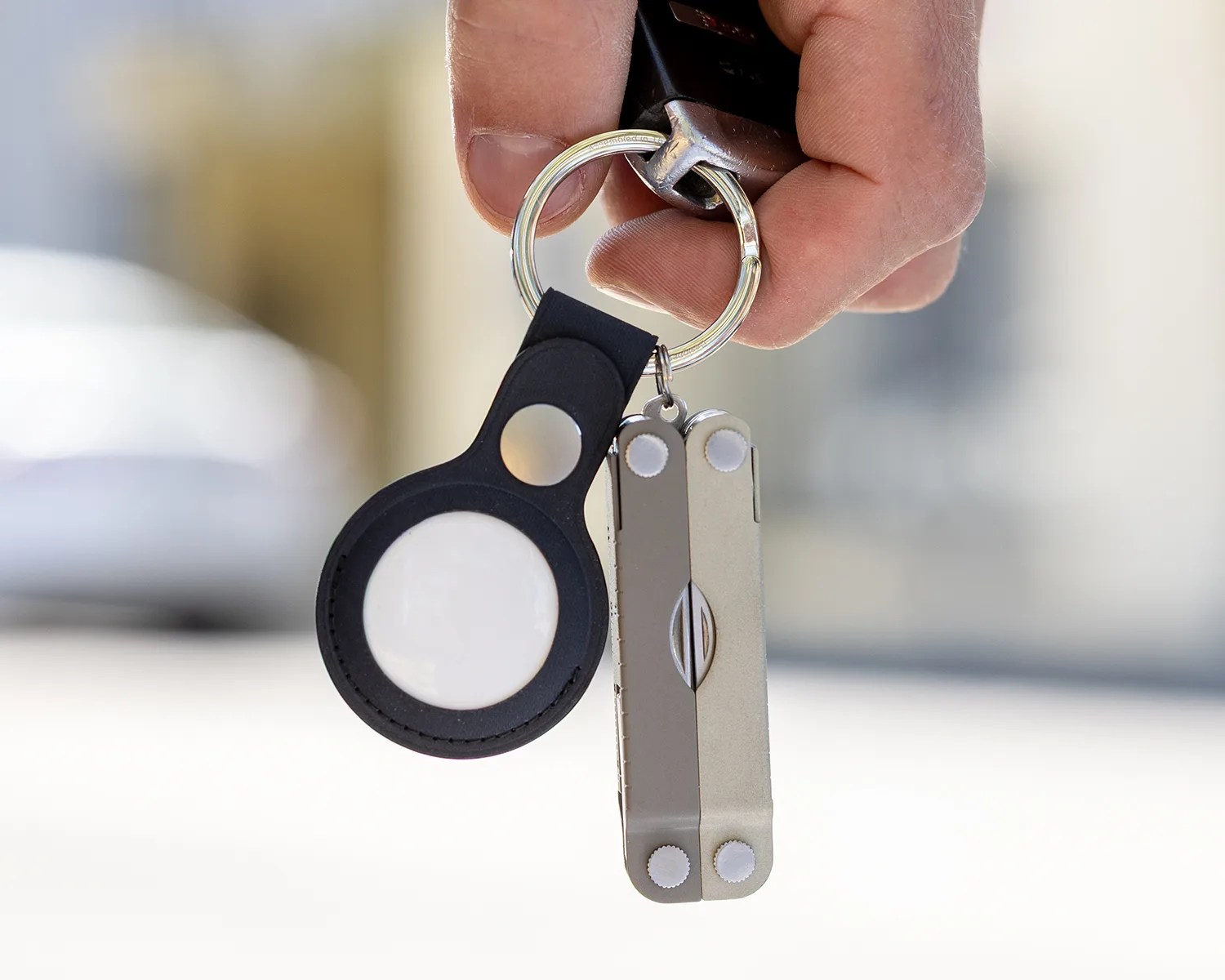 A hand holding a keyring with two items attached: a black AirTag holder with a white circular AirTag inside, and a compact silver multi-tool. The background is blurred, focusing on the keyring and hand.