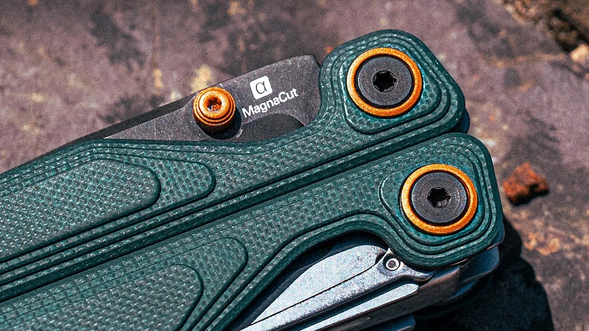 Close-up of a multitool with textured green handles and orange-accented pivot screws. The blade is partially visible and marked with "MagnaCut" and a small alpha symbol. The background is a rough, dark surface.