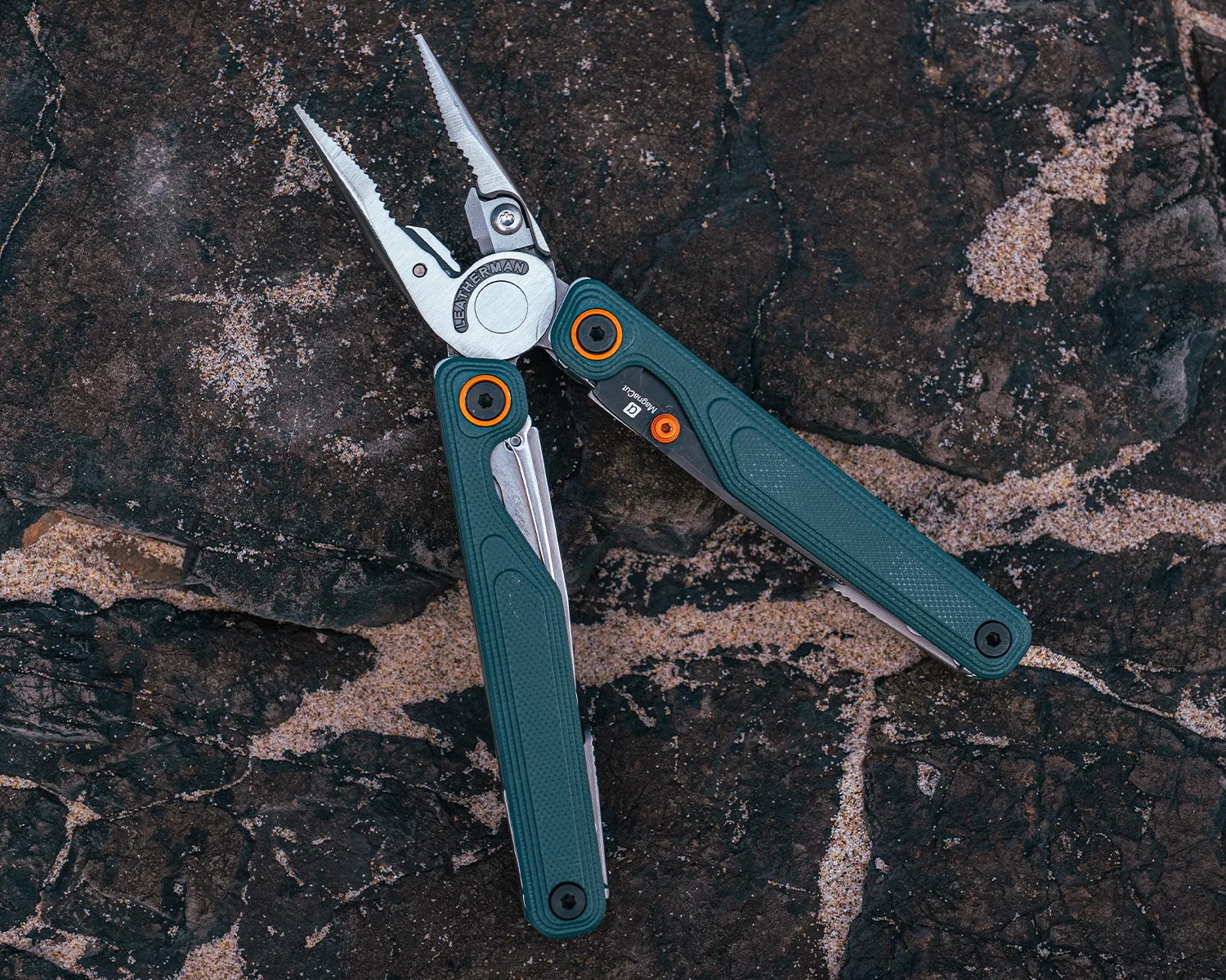 A Leatherman multi-tool with green textured handles and orange accents, featuring needle-nose pliers, is open and placed on a dark, textured rock surface. The tool's metal parts are silver, and the brand name "Leatherman" is visible on the pivot joint.