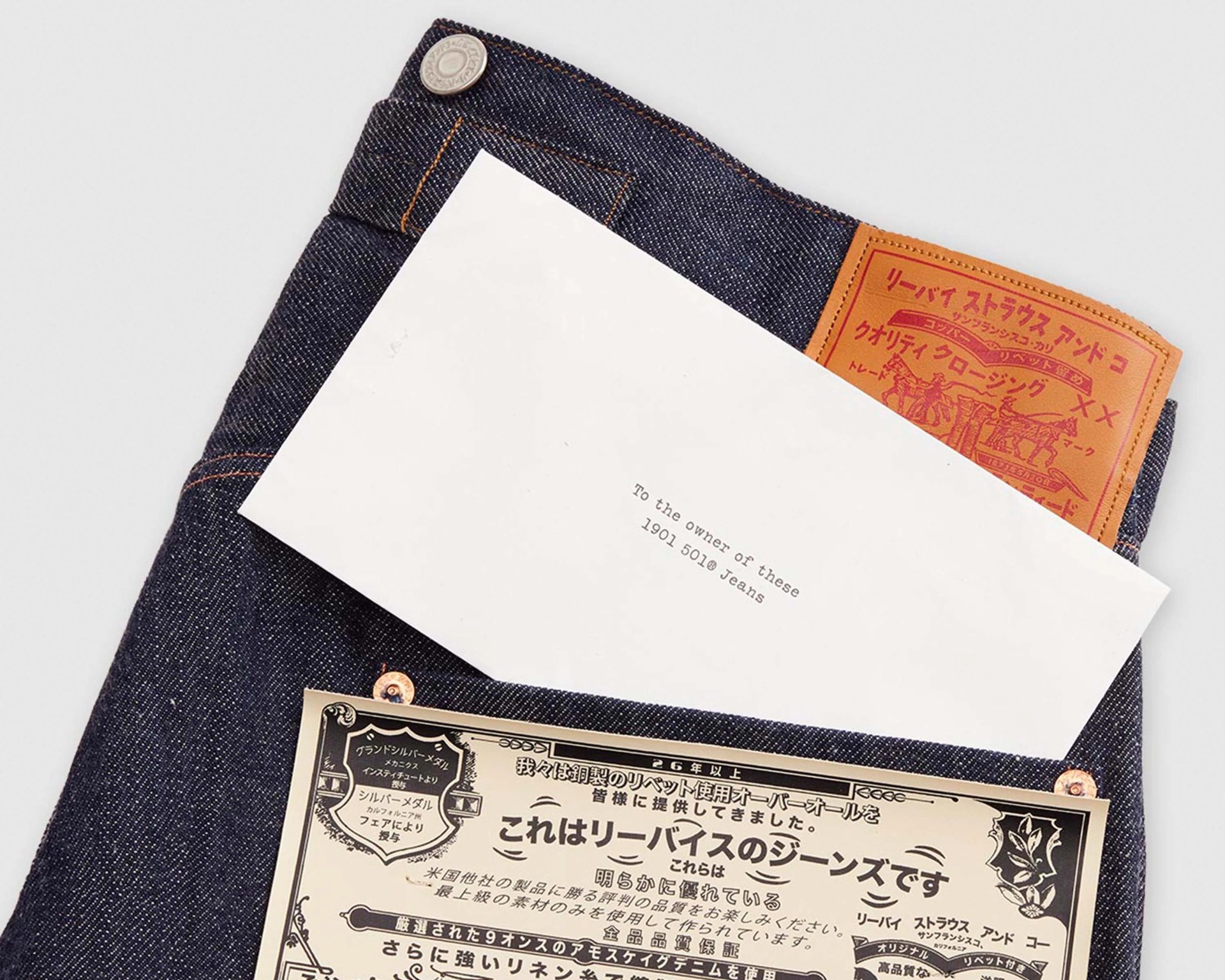 Dark blue denim jeans with a brown leather patch featuring red Japanese text and illustrations. A white envelope with the text "To the owner of these 1901 501® Jeans" is tucked into the waistband. A cream-colored label with Japanese text and decorative borders is attached to the jeans.