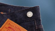 Close-up of the waistband of dark blue denim jeans with orange stitching, featuring a metal button with Japanese characters and a tan leather patch with red Japanese text.