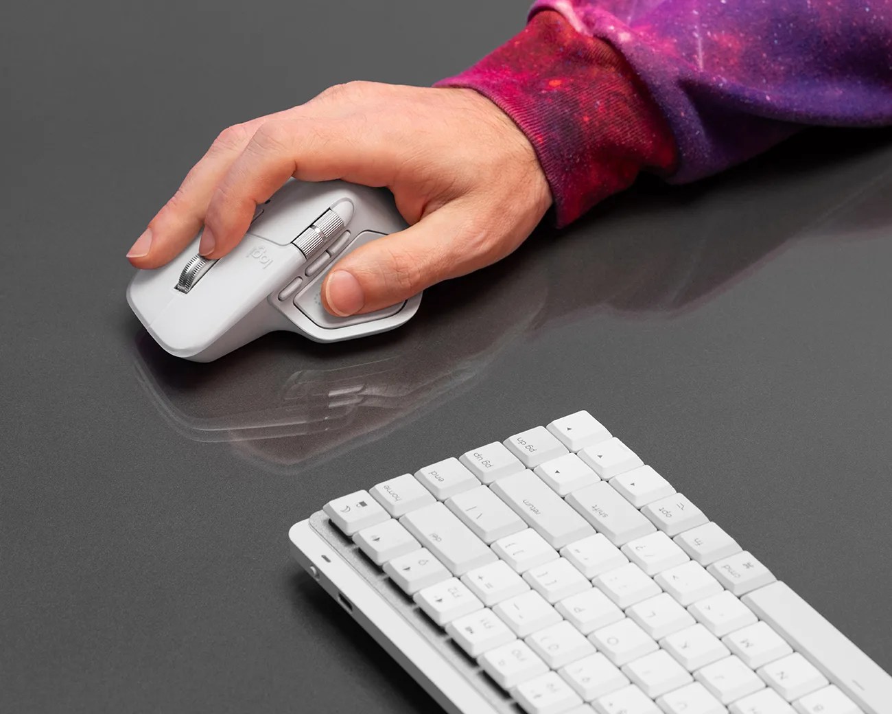 A hand wearing a purple and red long-sleeve shirt is using a white ergonomic computer mouse on a dark gray surface. A white keyboard is partially visible in the foreground.