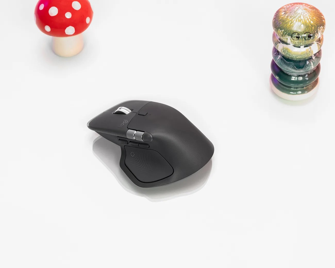 Black ergonomic computer mouse with silver scroll wheels, placed on a white surface. To the left is a red mushroom-shaped object with white spots, and to the right is a multicolored, stacked glass object.