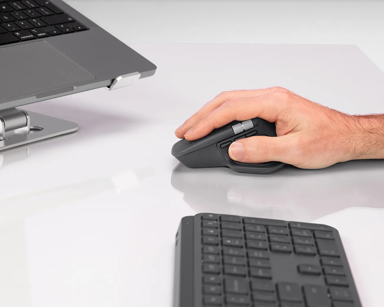 A right hand using a black ergonomic computer mouse on a white desk, with a partial view of a laptop on a stand and a black keyboard nearby.