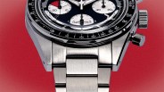 Close-up of a stainless steel wristwatch with a black bezel featuring white numerical markings. The watch face has three white subdials with black and red accents, and silver hands. The watch is set against a red background.