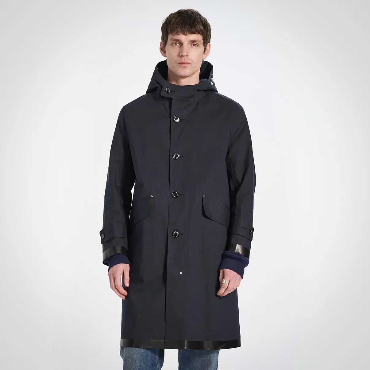 Navy blue long coat with a hood, featuring black buttons down the front, two front flap pockets, and black trim details on the cuffs and hem. The coat has a straight cut and is worn over a dark blue long-sleeve top and blue jeans.