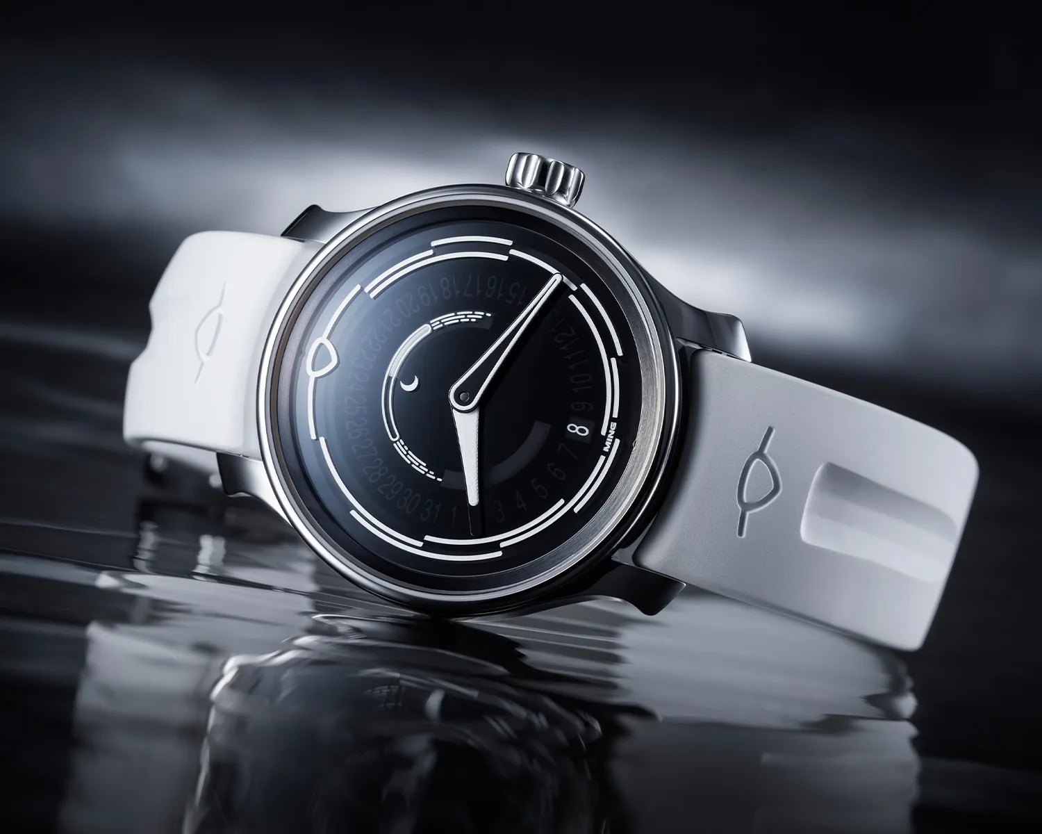 A sleek wristwatch with a black dial featuring white markings and numerals, encased in a polished silver case. The watch has two hands, one for hours and one for minutes, both in silver. It is paired with a white rubber strap embossed with a minimalist logo. The watch is positioned on a reflective surface with a dark, blurred background.
