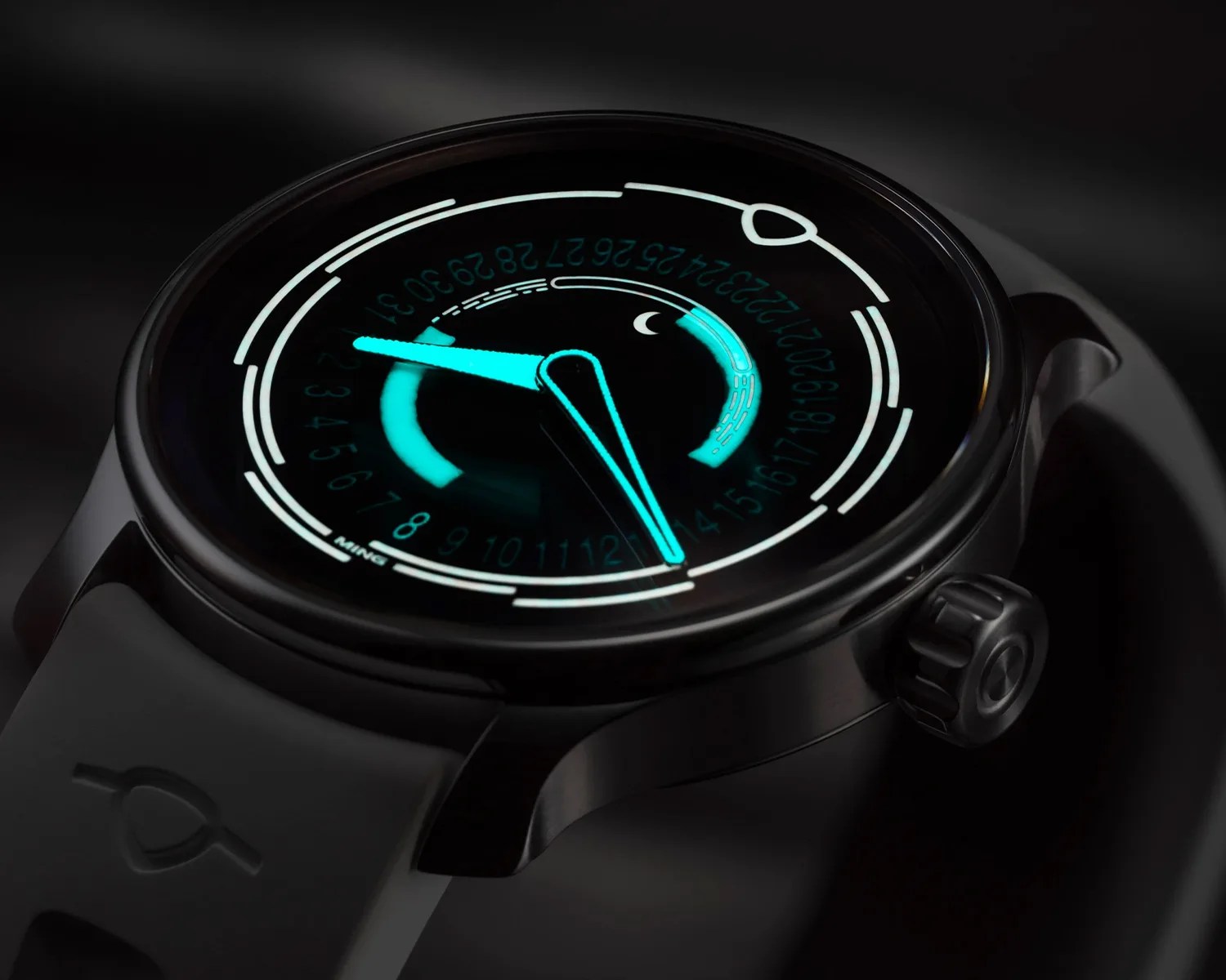 A close-up of a modern black wristwatch with a glowing blue display. The watch face features a circular design with numbers 1 to 31 around the inner edge, a crescent moon icon, and two prominent glowing blue hands indicating the time. The watch has a rubber strap with a subtle embossed logo near the lugs. The overall design is sleek and futuristic with luminous elements.