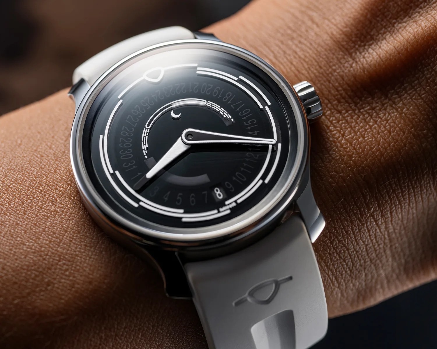 A close-up of a wristwatch with a black dial featuring concentric circular markings and numbers. The watch has two silver hands and a crescent moon symbol near the center. It is attached to a white strap with a subtle embossed logo. The watch is worn on a person's wrist.