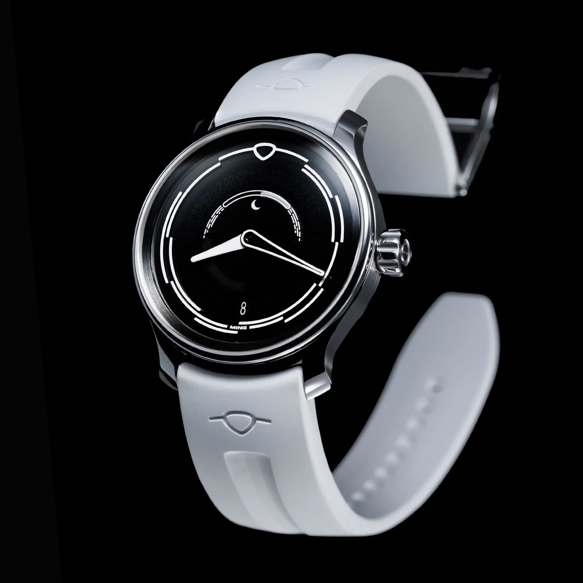 A sleek wristwatch with a polished silver case and a black dial featuring white minimalist markings and hands. The watch has a white rubber strap with a subtle embossed logo near the lugs. The dial includes a small crescent moon symbol and a date window at the 6 o'clock position, with the brand name "MING" displayed just above the date. The crown is textured for grip, and the overall design is modern and elegant against a black background.