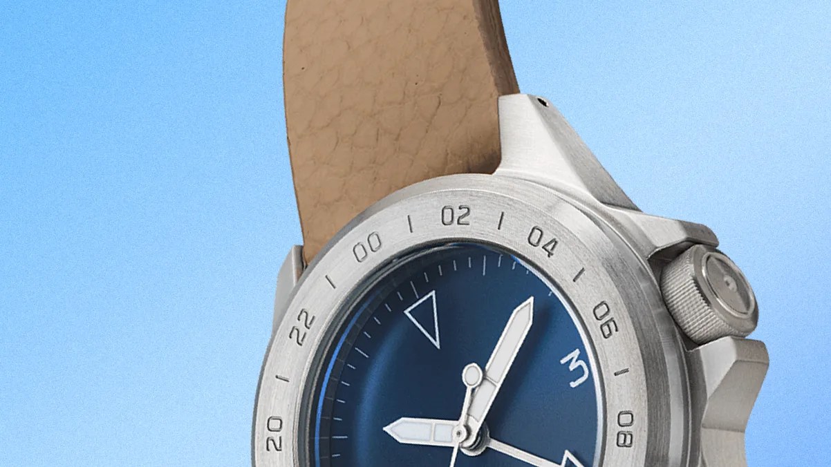Close-up of a wristwatch with a blue dial, silver bezel marked with 24-hour numerals, and a beige leather strap. The watch has silver hour and minute hands, with a triangular marker at the 12 o'clock position. The crown is textured for grip and positioned on the right side of the watch case. The background is a gradient light blue.