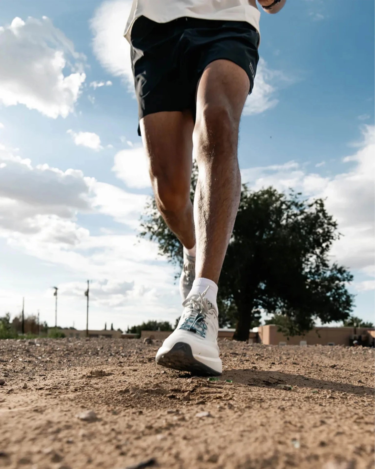 Close-up of a person running outdoors on a dirt path, wearing white running shoes, white socks, and black shorts, with a partly cloudy blue sky and a tree in the background.