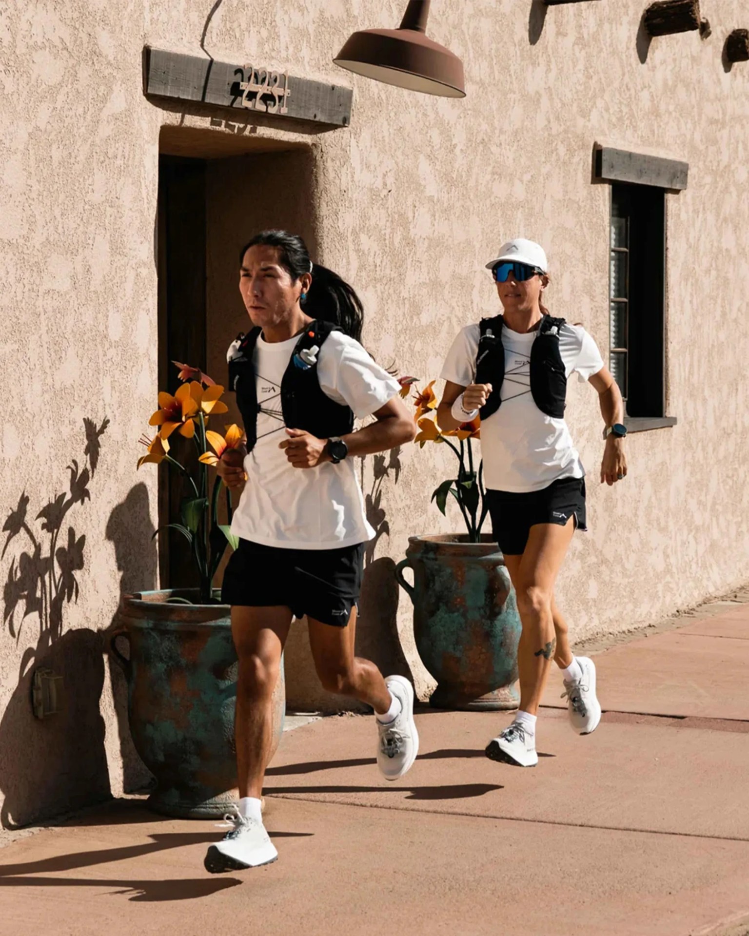 Two runners wearing white shirts, black shorts, black running vests, and white running shoes are jogging on a sunlit sidewalk next to a beige stucco wall with a door and window. One runner has long dark hair tied back, and the other wears a white cap and blue sunglasses. Large ceramic pots with orange flowers are placed along the wall.