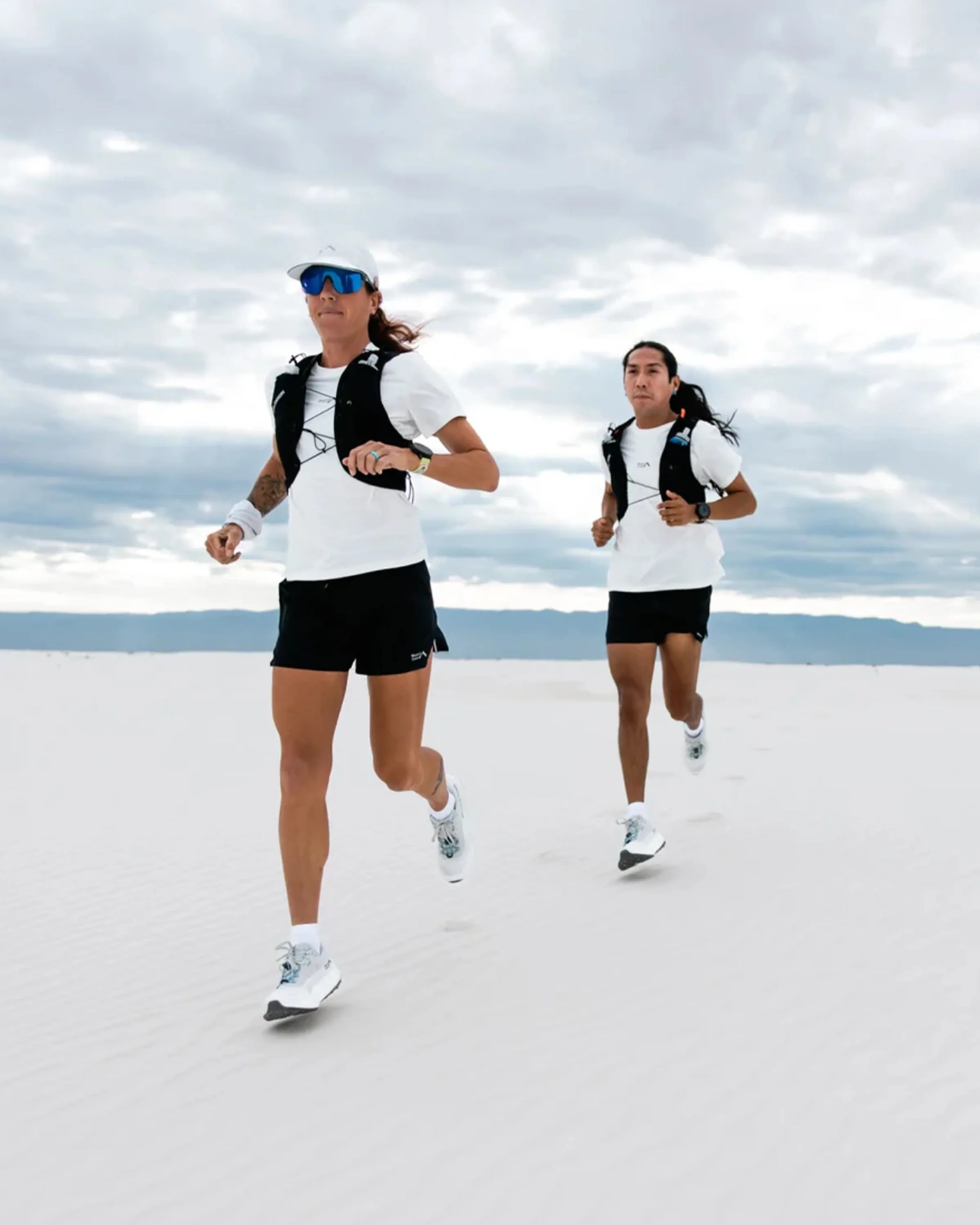 Two people are running on a white sandy surface under a cloudy sky. Both are wearing white t-shirts, black shorts, black running vests, and white running shoes. The person in front is also wearing a white cap and blue reflective sunglasses.