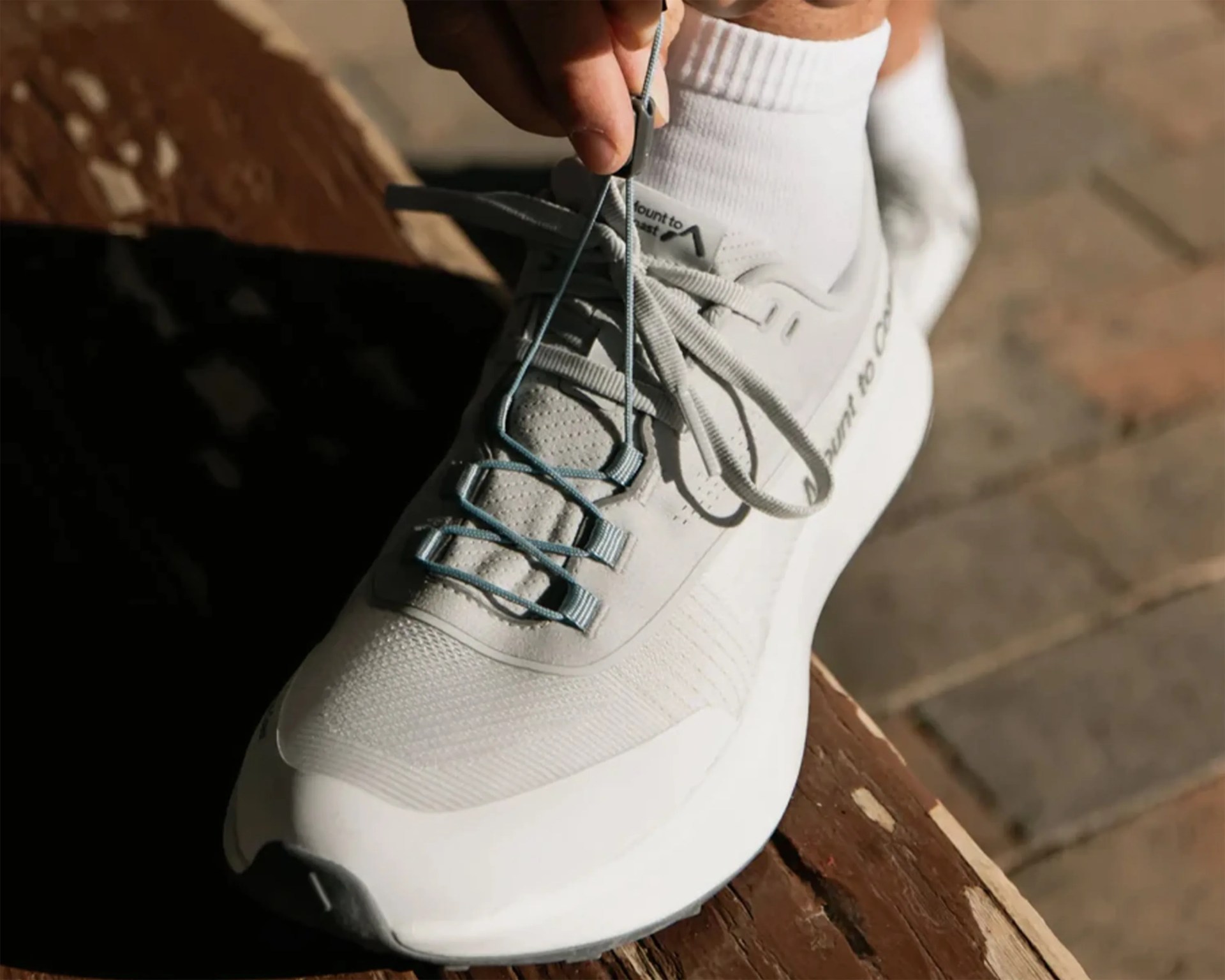 A close-up of a white athletic shoe being laced up by a person wearing white socks. The shoe has a textured mesh upper with light blue lace loops and a thick white sole. The shoe is resting on a wooden surface with a blurred stone or tile floor in the background.