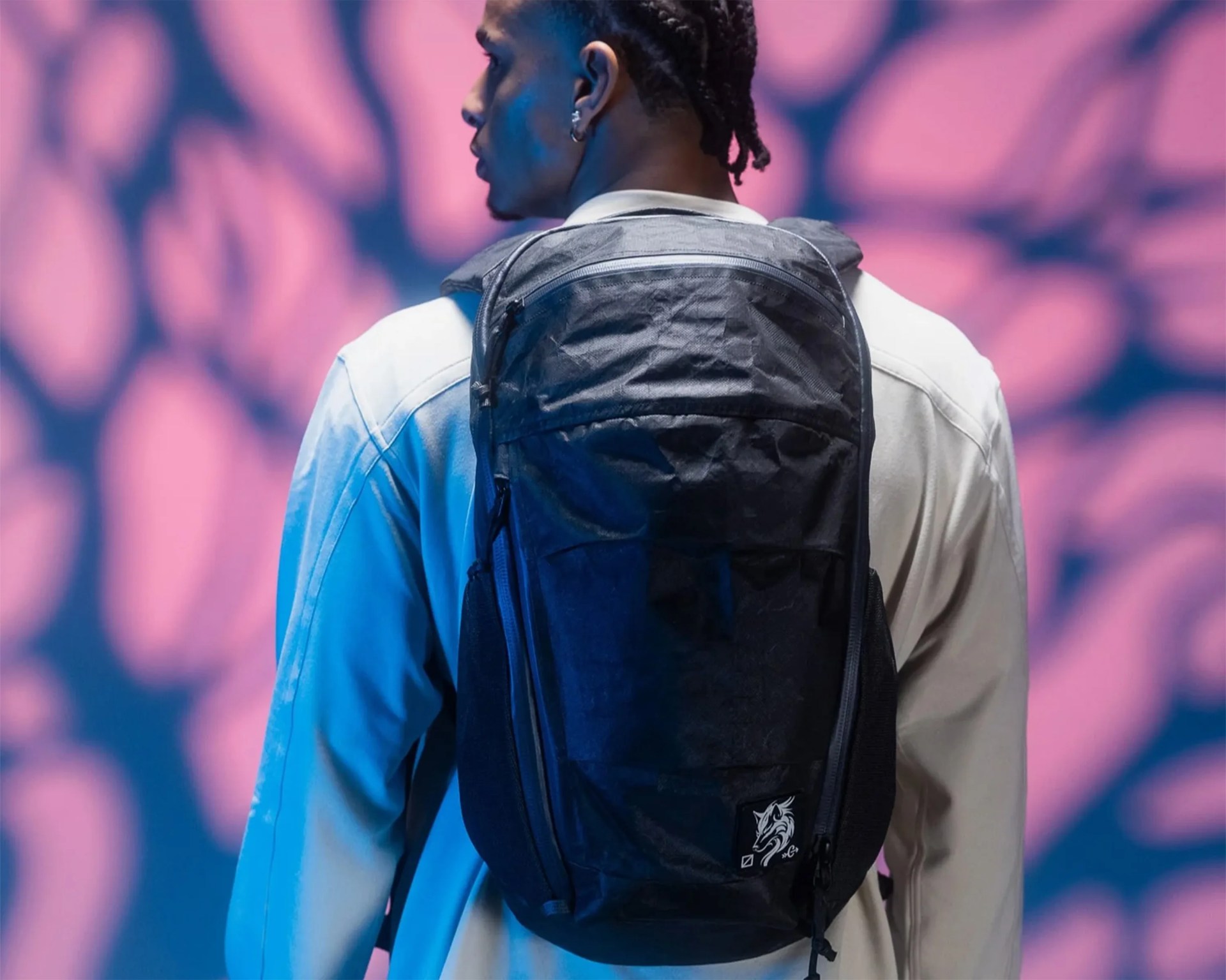 A person wearing a light-colored jacket and a black backpack with a small white dragon logo on the lower right corner. The background features a pink and blue abstract pattern.