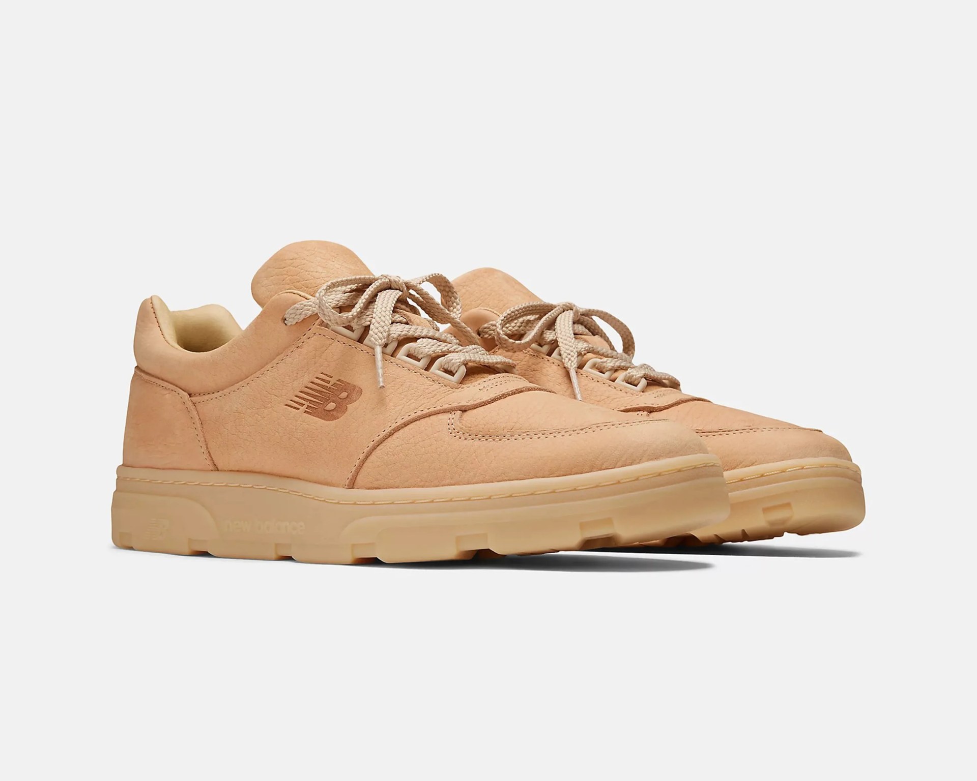 Pair of beige New Balance sneakers with a textured leather upper, matching beige laces, and a thick rubber sole. The New Balance logo is embossed on the side of each shoe. The design is minimal and monochromatic, with a slightly padded collar for comfort.