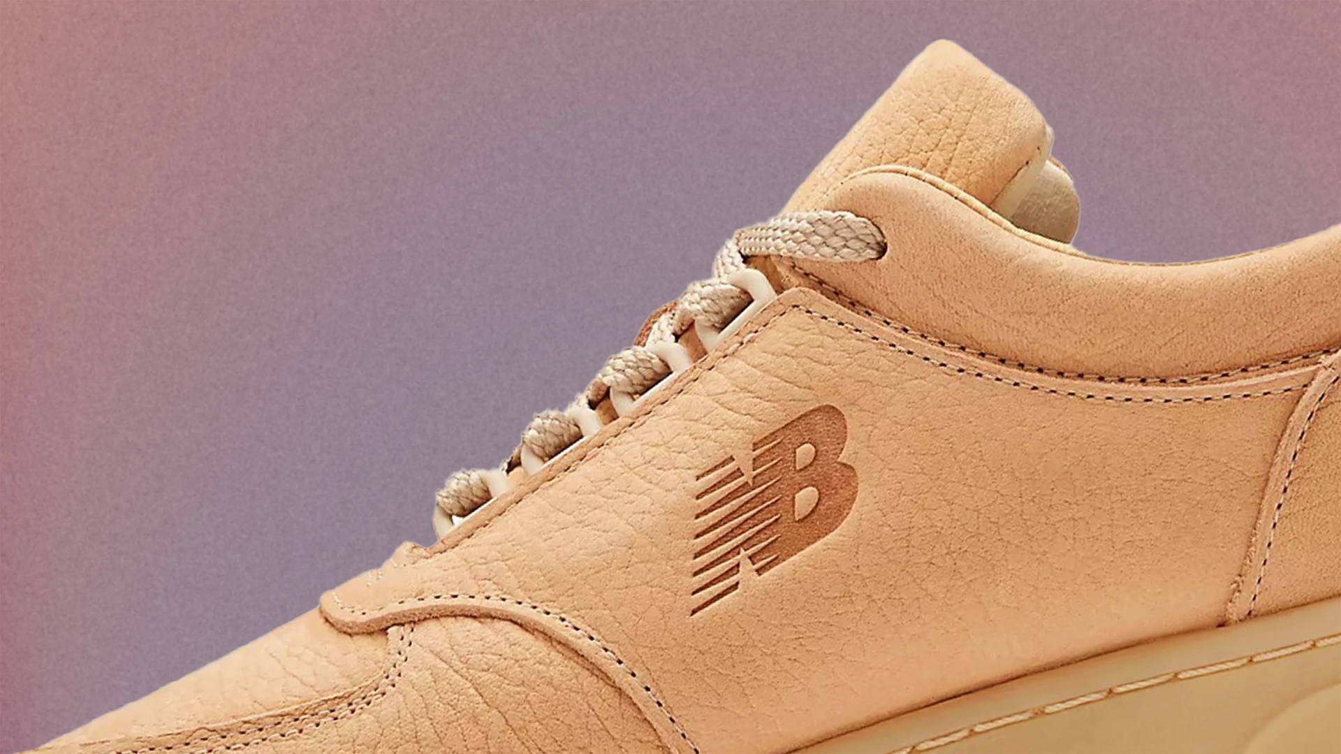 Close-up of a beige New Balance sneaker showing the textured leather upper, beige laces, and the embossed New Balance "NB" logo on the side. The shoe has detailed stitching and a padded collar.