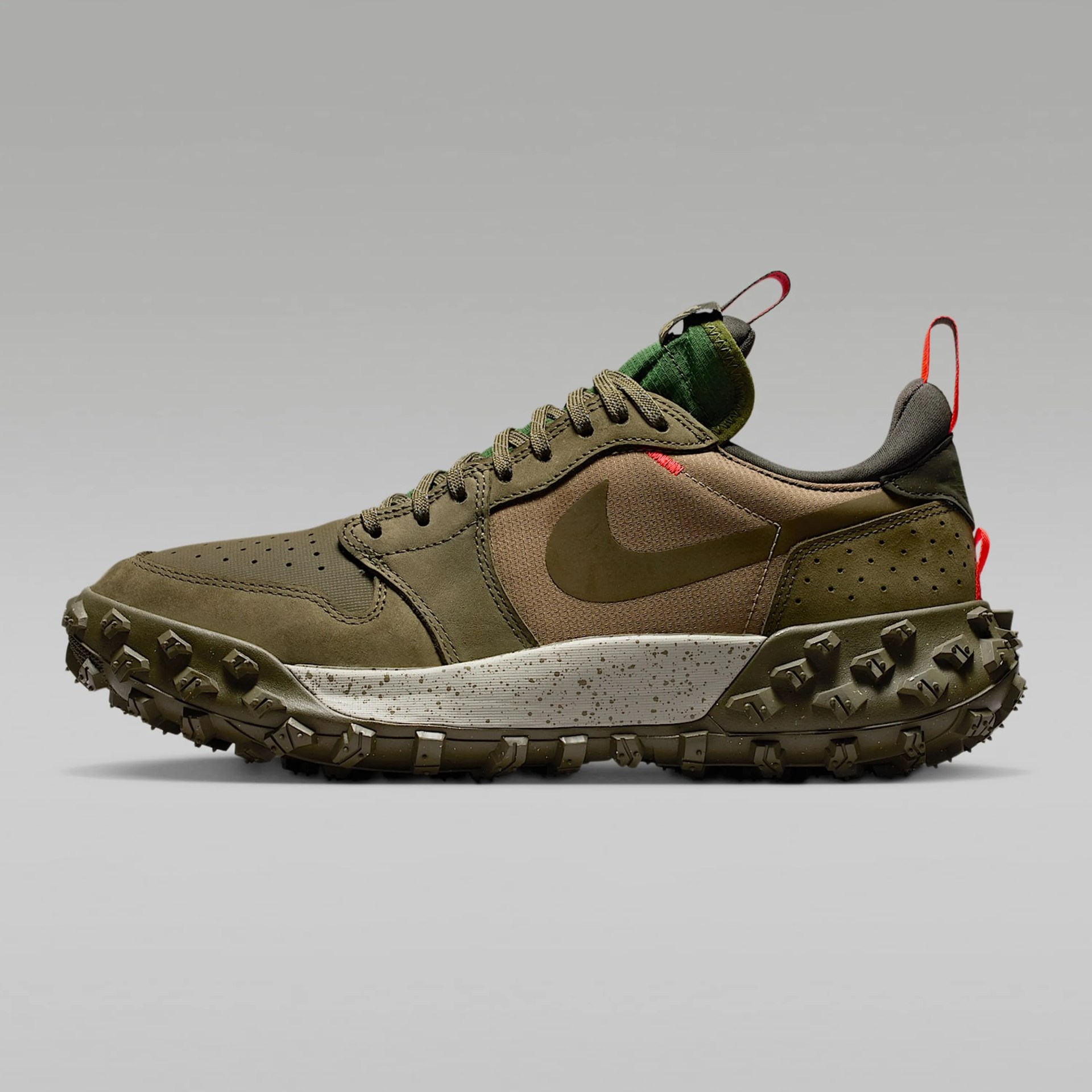Olive green and brown Nike sneaker with a rugged, heavily textured sole featuring large, angular treads. The shoe has a speckled white midsole, green laces, and red pull tabs on the tongue and heel. The Nike swoosh is prominently displayed on the side in a matching olive green color. The upper combines suede and mesh materials for durability and breathability.