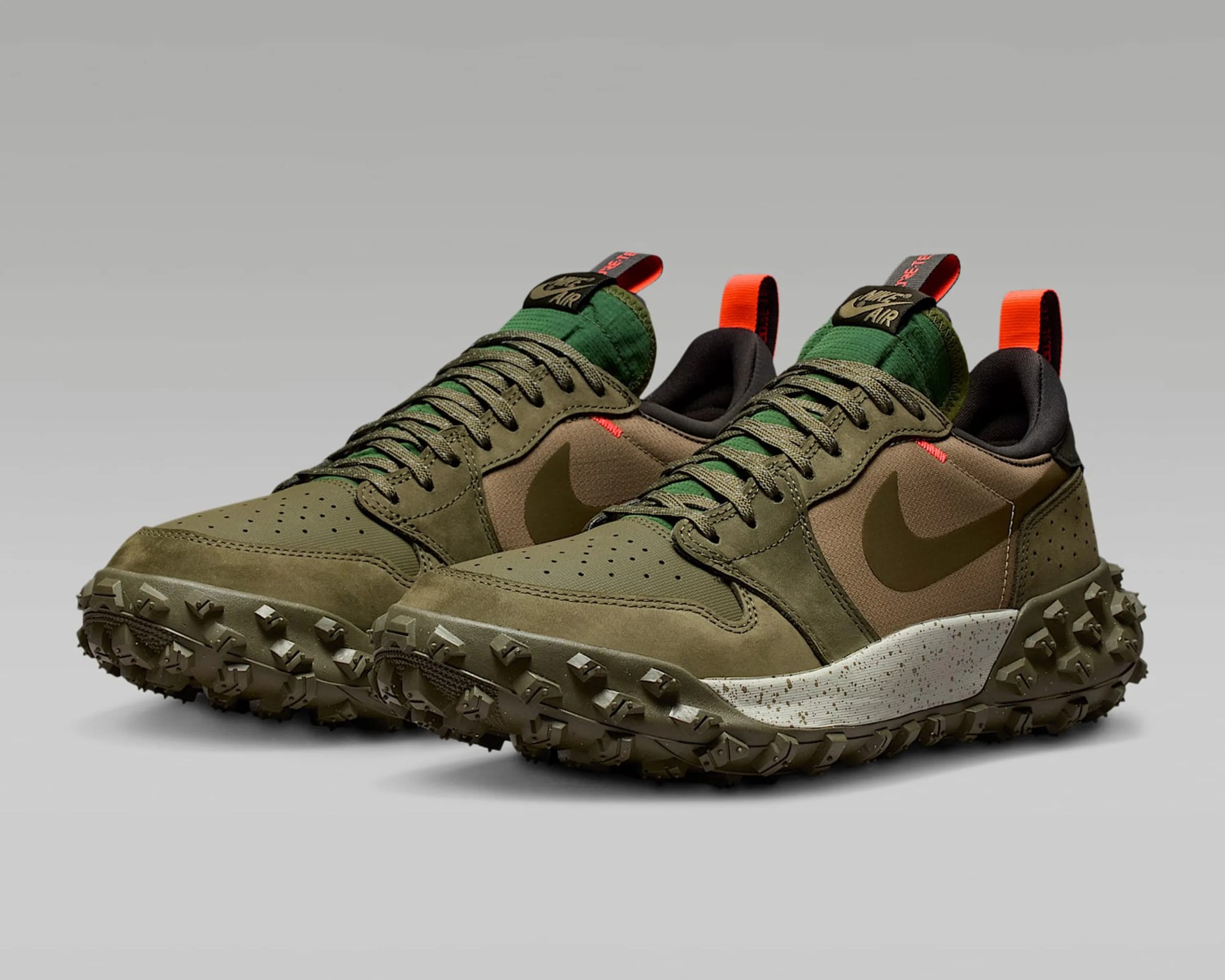 A pair of Nike sneakers in olive green and brown tones with a rugged, textured sole designed for traction. The shoes feature green laces, a green tongue with a black and gold Nike Air label, and bright orange pull tabs at the heel. The midsole is white with speckled detailing, and the Nike swoosh is prominently displayed on the side in a darker olive shade. The overall design combines outdoor durability with sporty style.