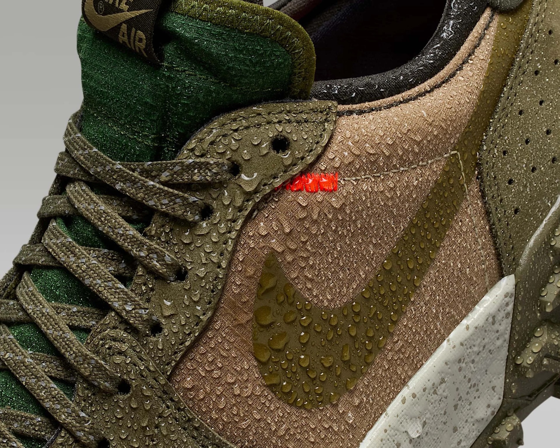 Close-up of a Nike sneaker featuring olive green and brown tones with water droplets on the surface. The shoe has textured fabric and suede materials, olive green laces, a green tongue with a Nike Air logo, and a brown Nike swoosh. The midsole is white with speckled detailing.