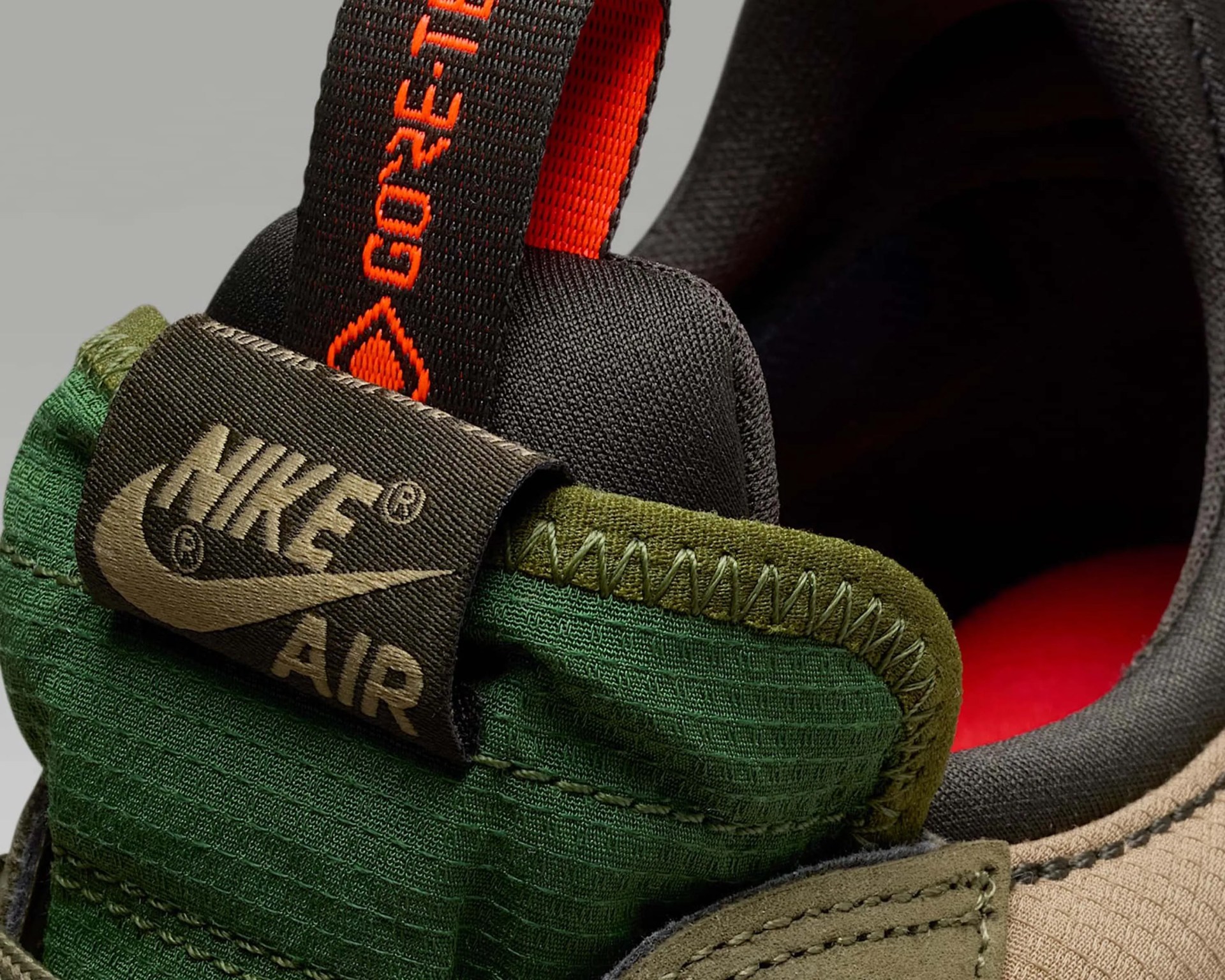 Close-up of the back heel area of a Nike Air shoe featuring green fabric with a textured pattern, olive green stitching, and a black pull tab with orange "GORE-TEX" text. The Nike Air logo is prominently displayed on a black fabric tag. The shoe's interior lining is black with a red insole visible inside.