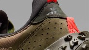 Close-up of the heel section of an olive green and brown sneaker with perforated suede material, black fabric lining, and a bright red pull tab. The shoe features a textured sole with rugged treads and a subtle embossed logo on the upper heel area.