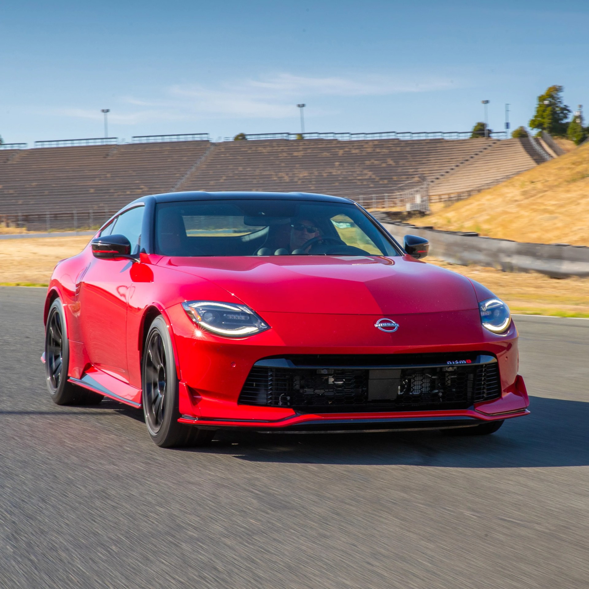 Red Nissan sports car with black roof and black wheels driving on a racetrack, with empty bleachers and dry grass in the background.