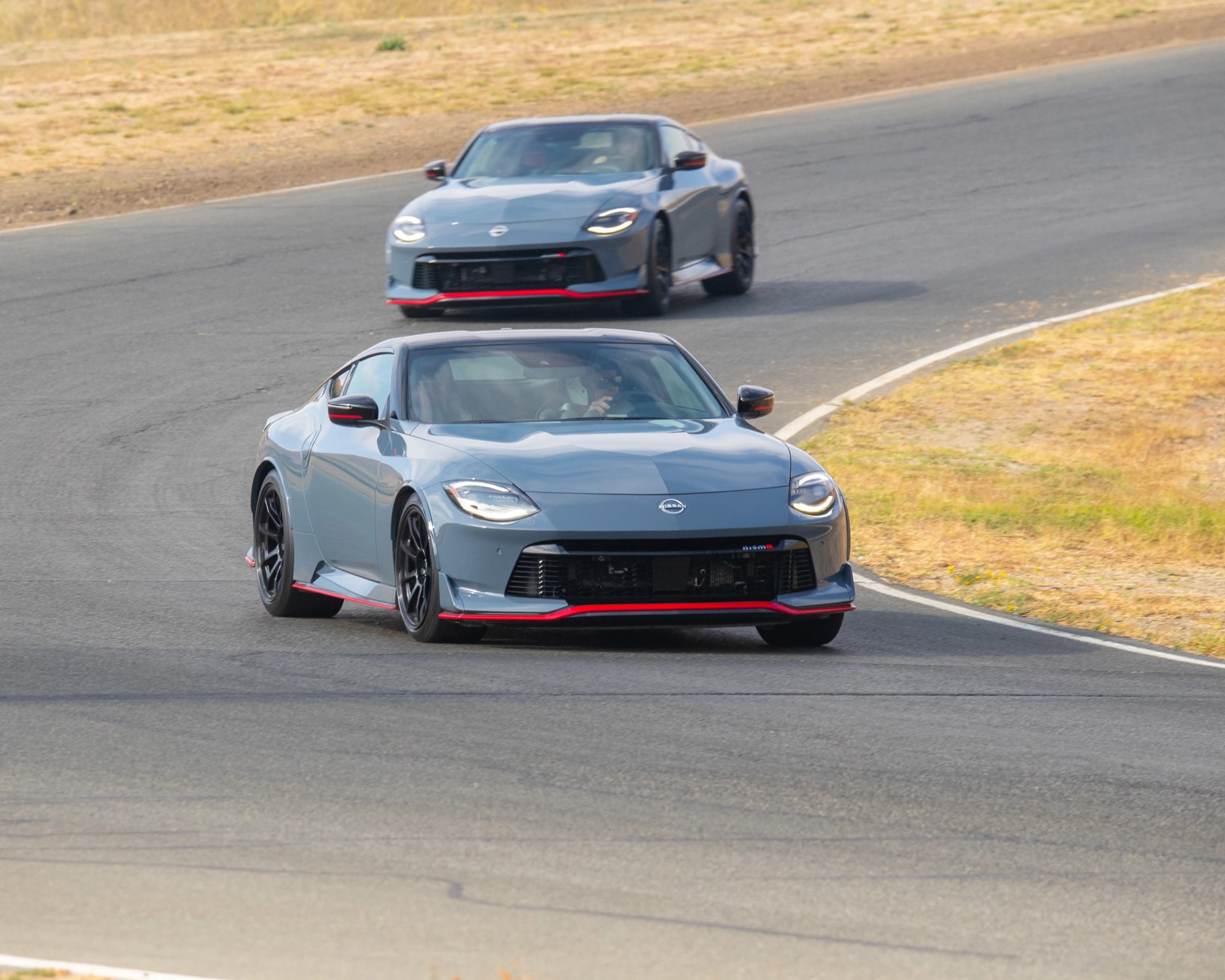 Two gray Nissan sports cars with black wheels and red accents are driving on a curved race track. The cars have sleek, aerodynamic designs with prominent front grilles and LED headlights. The track is surrounded by dry grass and dirt.