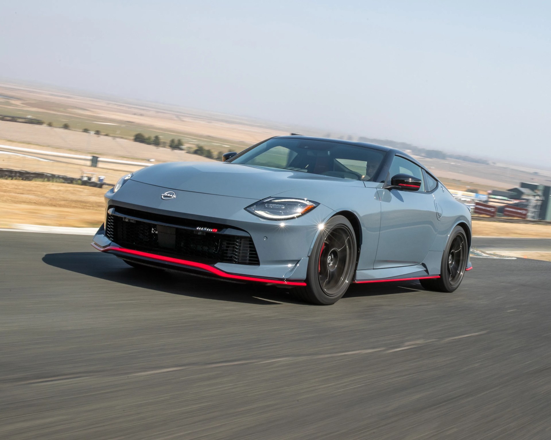 Gray Nissan sports car with black wheels and red accents on the front lip and side skirts, driving on a racetrack with a dry, open landscape in the background.