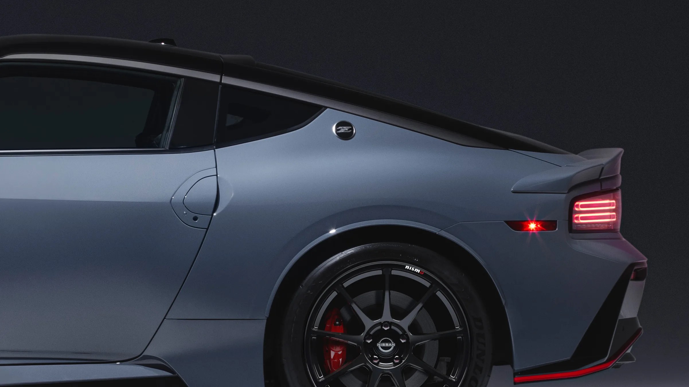Rear side view of a sleek, modern sports car in matte gray with a black roof. The car features a distinctive "Z" emblem near the rear window, black multi-spoke alloy wheels with red brake calipers, and illuminated horizontal rear lights. The design includes a subtle rear spoiler and aerodynamic contours.