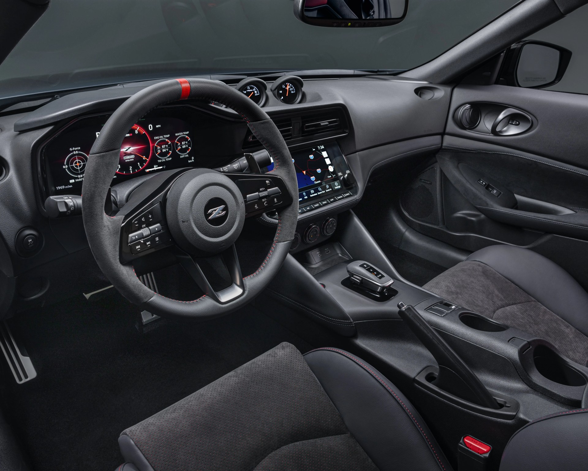 Interior of a modern sports car featuring a black leather and suede steering wheel with a red center stripe and a "Z" emblem in the center. The dashboard includes a digital instrument cluster displaying various gauges and a central touchscreen infotainment system showing a navigation map. The center console has a compact gear selector, manual handbrake, cup holders, and climate control knobs. The seats are upholstered in a combination of black leather and suede with red stitching accents. The door panel includes a speaker, window controls, and a door handle.
