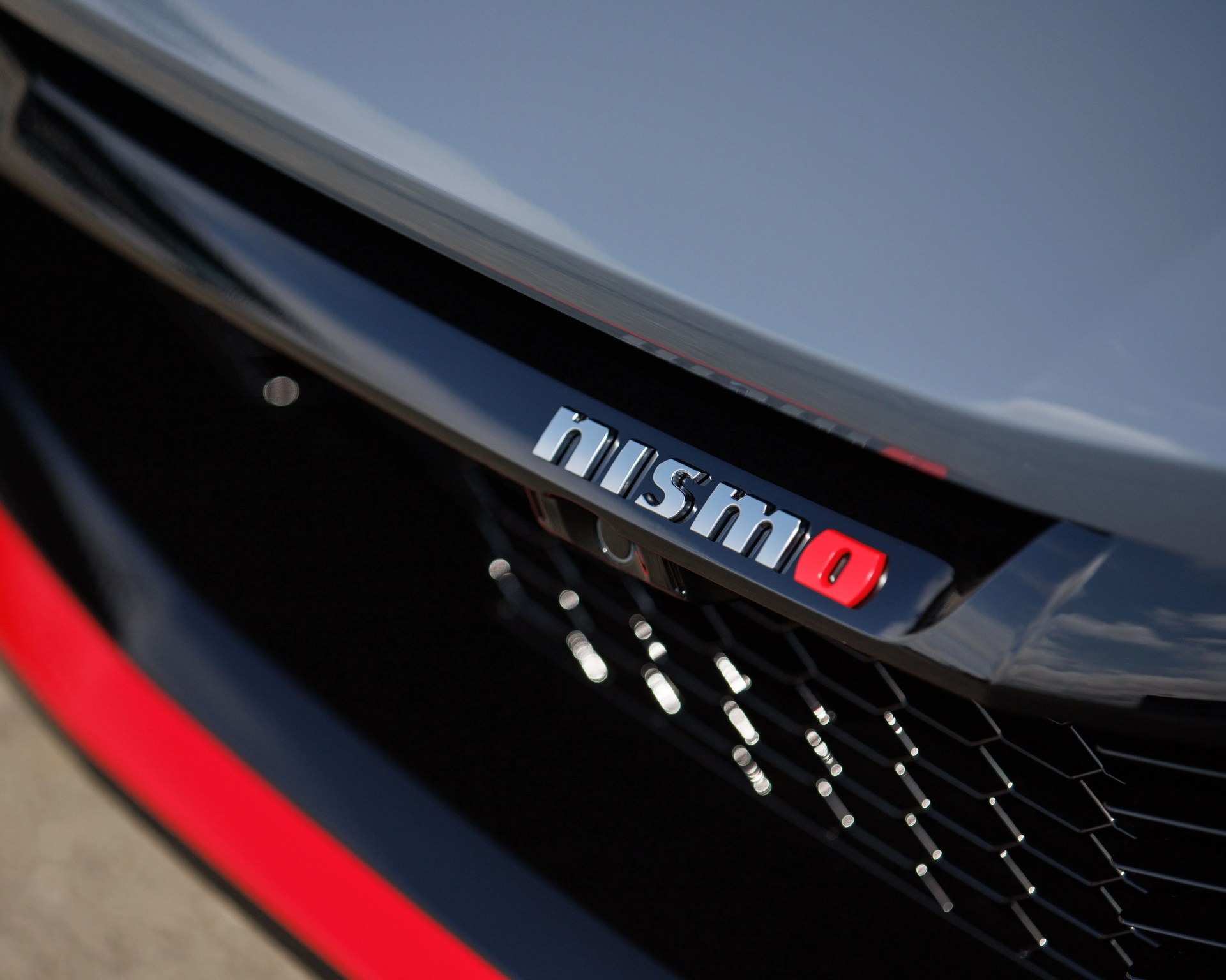 Close-up of a car grille featuring a "nismo" badge with silver letters and a red "o" on a black background, with part of the car's gray body and a red accent visible.
