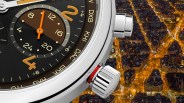 Close-up of a luxury watch face showing a 24-hour subdial with orange hands and gold numerals, encased in polished silver metal. The watch bezel features city abbreviations like DXB, KHI, DAC, and BKK, indicating world time zones. The crown and a push button are visible on the right side of the watch case. The background shows a nighttime aerial view of a city with illuminated streets and buildings.
