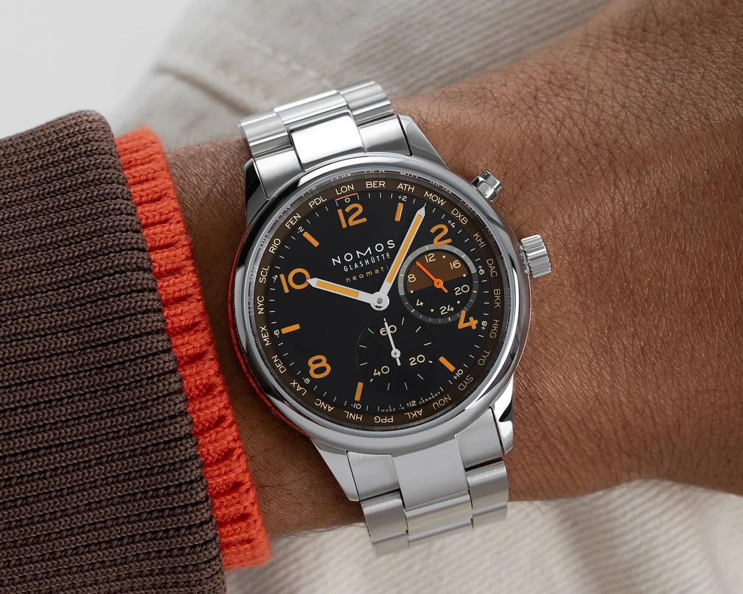 Silver wristwatch with a black dial featuring orange Arabic numerals and markers, two subdials, and white and orange hands. The watch has a stainless steel bracelet and is worn on a wrist with brown skin, paired with a brown and orange knit sleeve. The brand "NOMOS GLASHÜTTE" and the word "neomatik" are visible on the dial.
