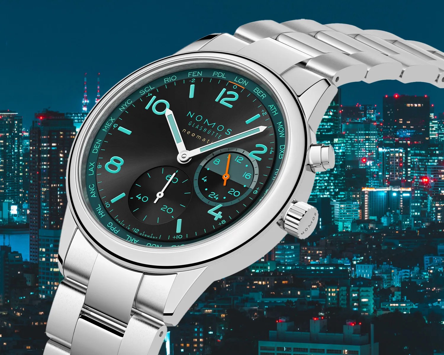 Silver metal wristwatch with a black dial featuring turquoise numerals and markers. The watch has two subdials, one for seconds and one for 24-hour time, with an orange hand on the 24-hour subdial. The bezel is smooth and polished, and the watch has a matching silver metal link bracelet. The brand name "NOMOS Glashütte" and the model "neomatik" are printed on the dial. The background shows a cityscape at night with illuminated buildings.