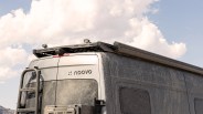 The rear upper section of a dusty gray van with a roof rack, two small mounted lights, a ladder on the left side, and the brand name "noovo" visible on the back door. The sky is partly cloudy in the background.