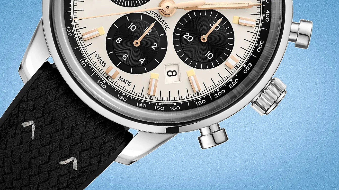 Close-up of a wristwatch featuring a steel stainless steel case, a cream-colored dial with two black subdials, and a date window at the 6 o'clock position. The watch has gold-tone hour markers and hands, a black tachymeter scale on the outer edge, and a black textured strap with white stitching. The crown and two pushers are visible on the right side of the case.