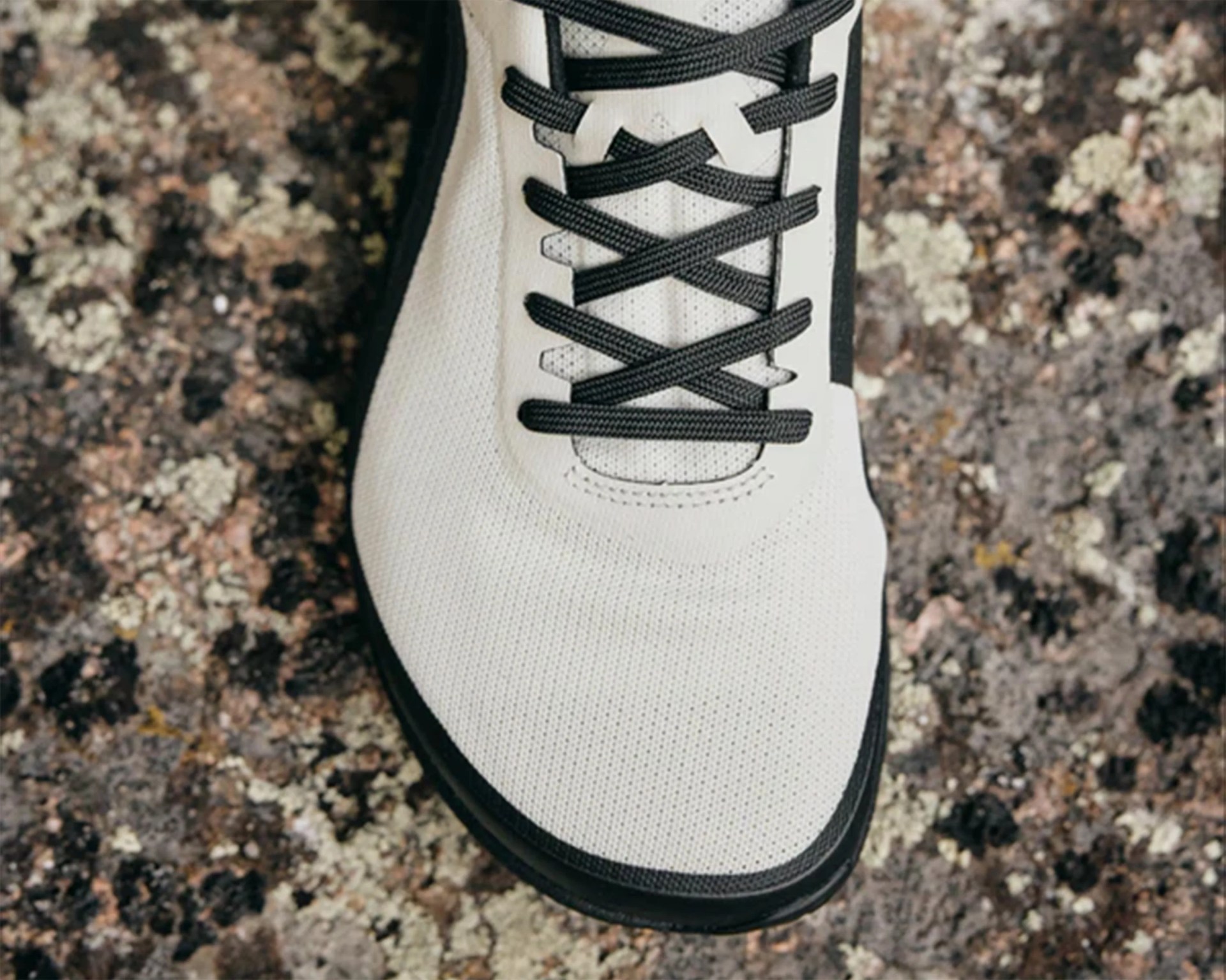 Close-up of the front part of a white sneaker with black laces and a black sole, resting on a textured, multicolored stone surface. The sneaker has a perforated design on the upper material.