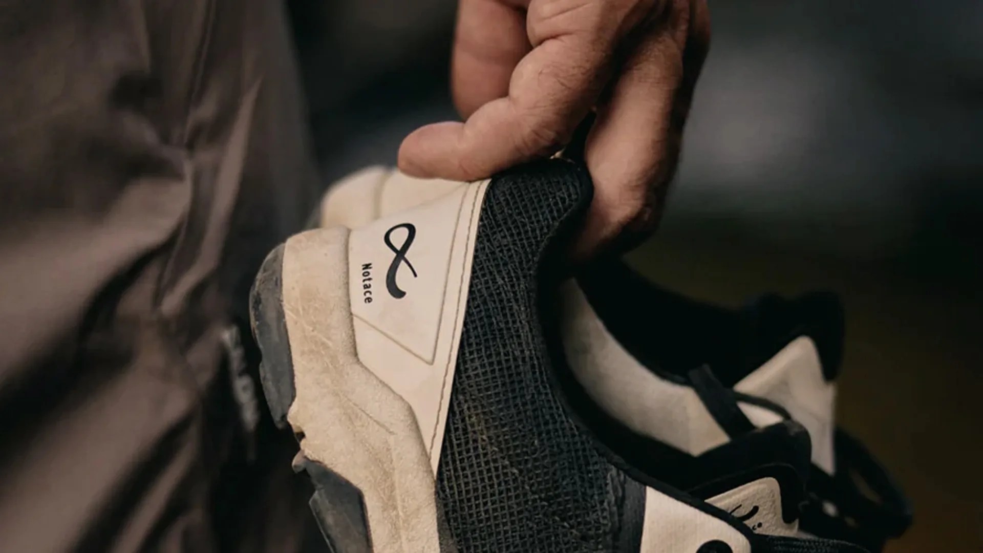 A close-up of a hand holding a black and white sneaker with a textured black mesh and a white heel area featuring a logo and the word "Notace." The sneaker appears slightly worn, and the background is blurred.